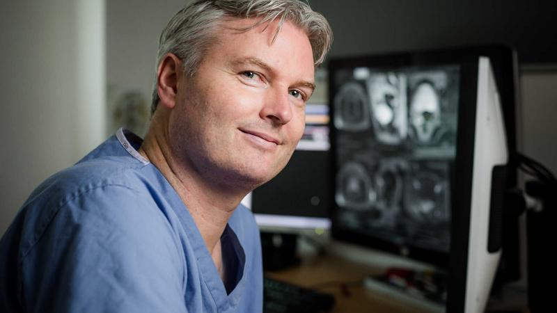 Radiologist smiling at his desk with mri scans displaying on the screen behind him