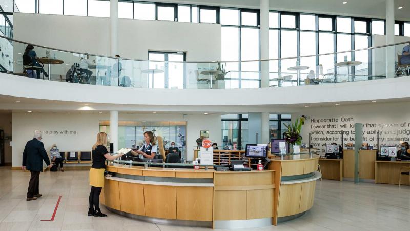 Reception desk with a woman handing paper to a patient while visitors walk around and sit on chairs,
